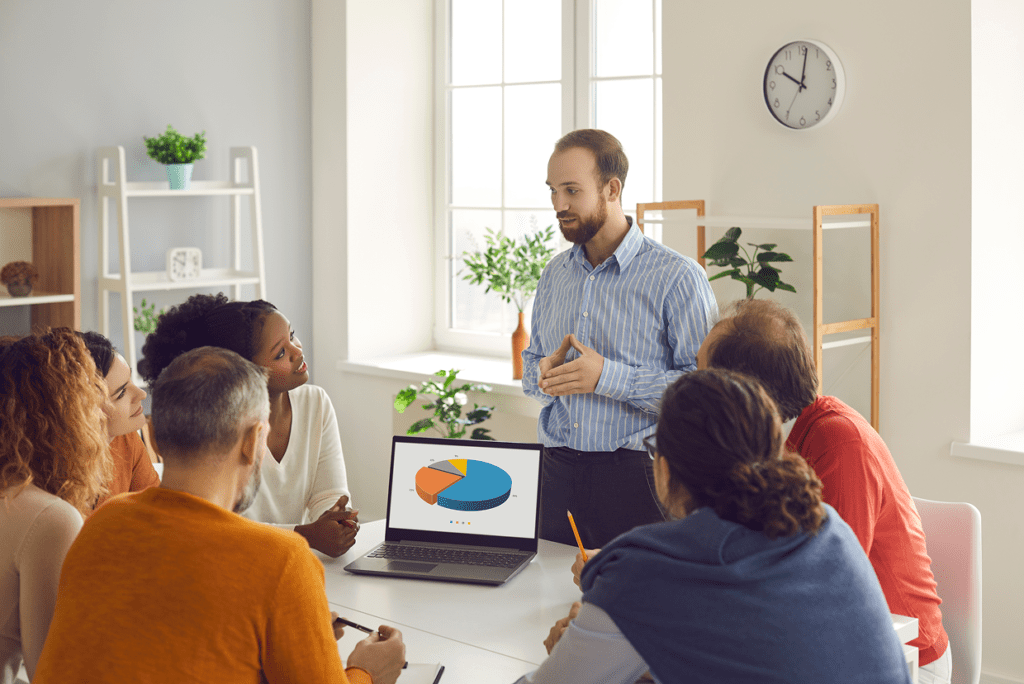 Man standing and making a presentation to a group of individuals seated around a desk