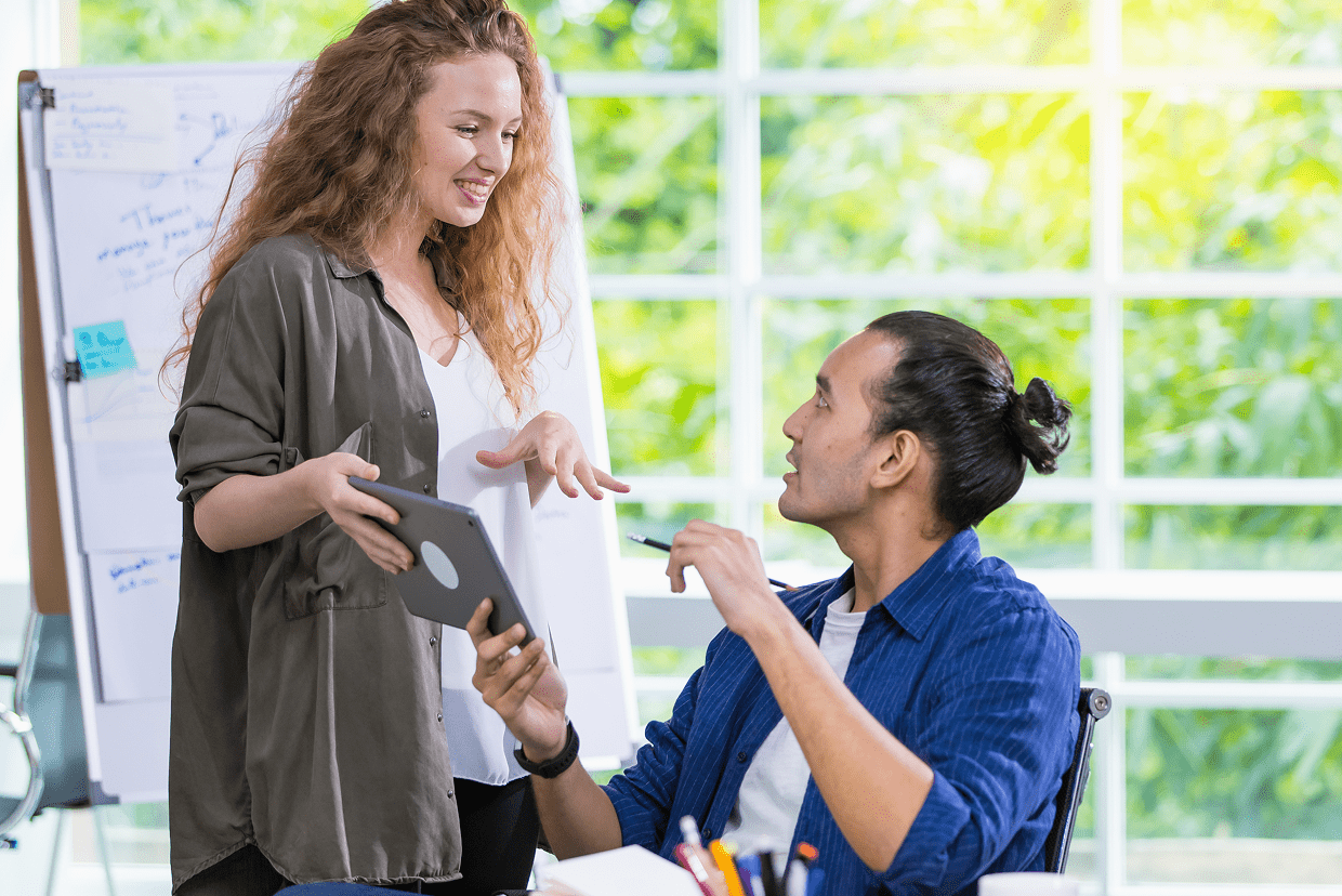 Female standing holding a tablet while talking to a make seated in a chair