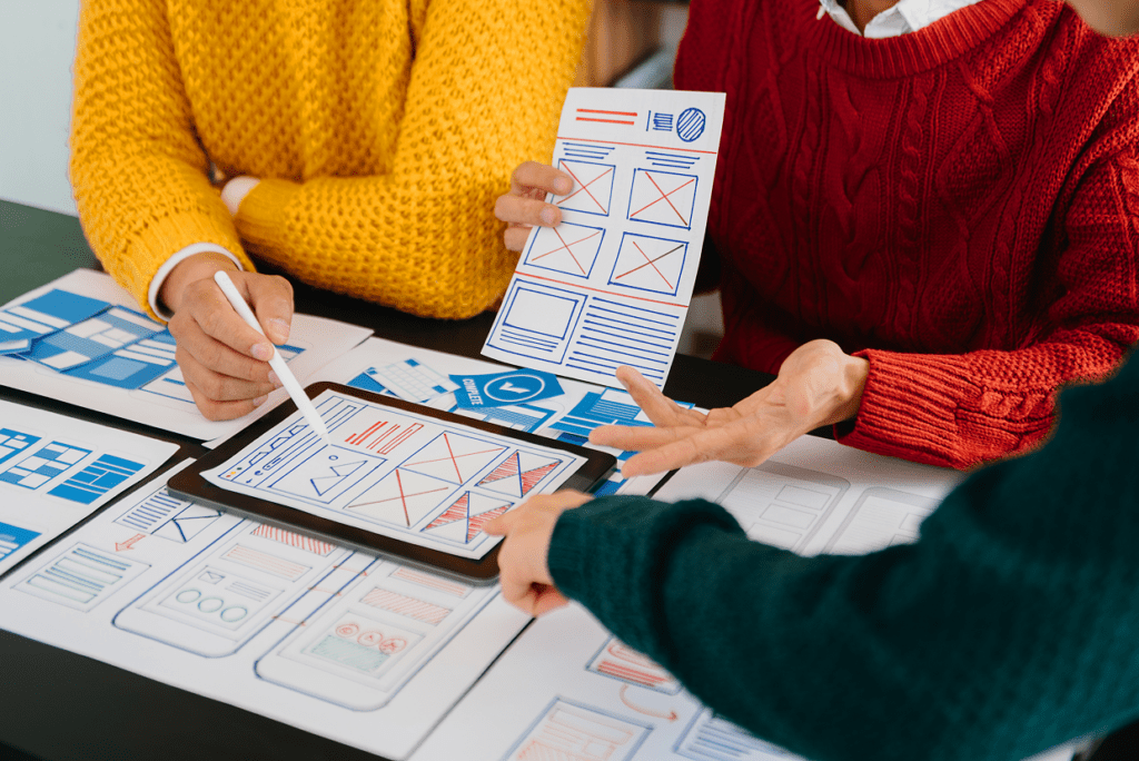 3 individuals each in a red, yellow, and green sweater seated around a table with multiple paper mockups