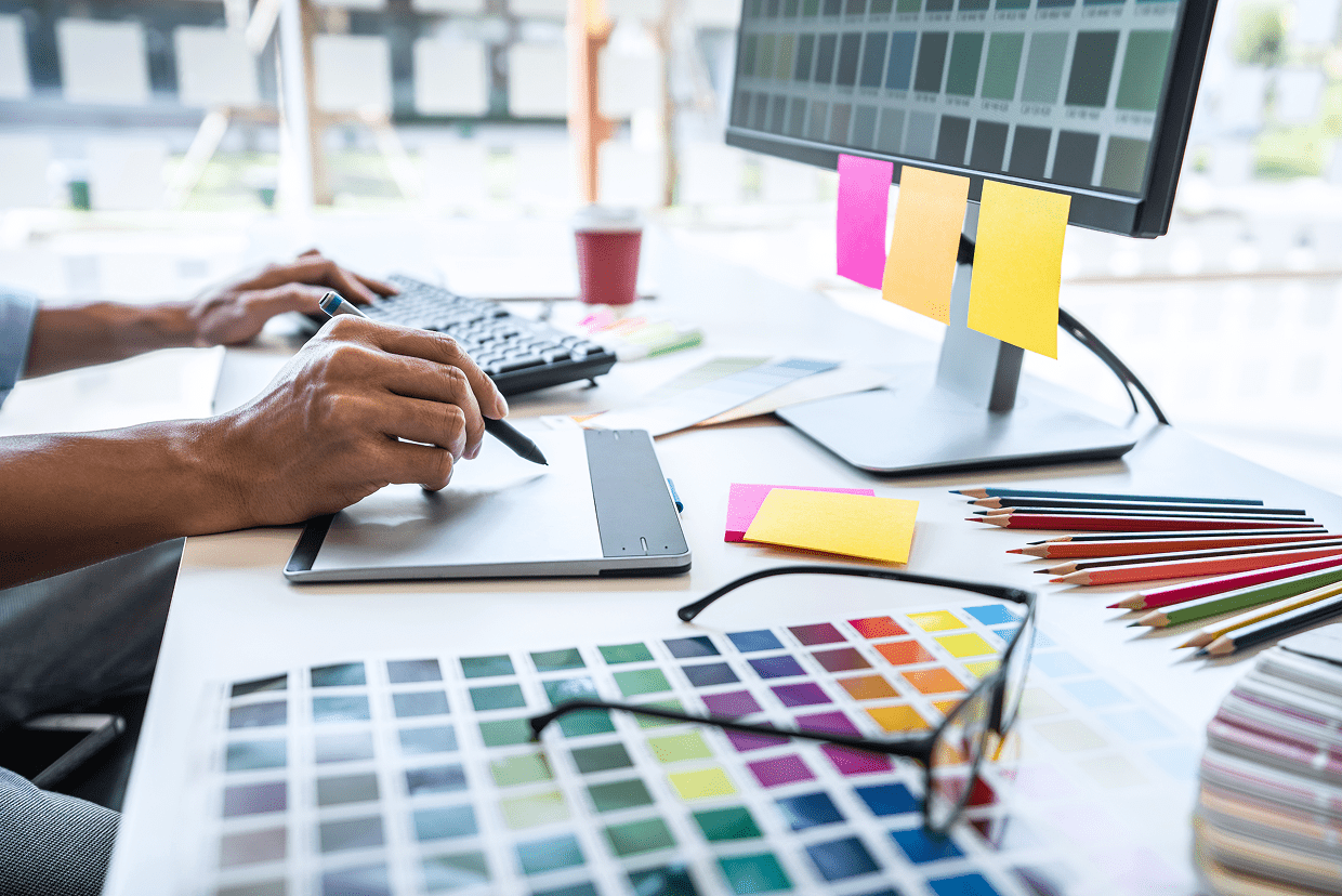 Male with left hand on keyboard and right hand with pen on mouse seated around a table with multiple color kits, pencils and post its