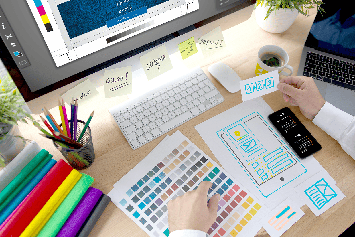 Light brown desk with multiple mockups, laptop, desktop monitor, and hand holding POSTIT note