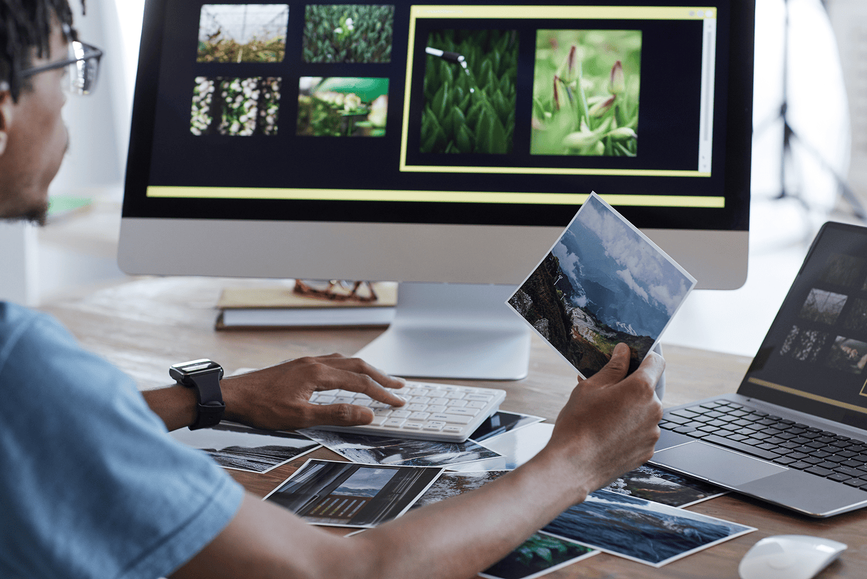 Man wearing a glasses looking at a monitor with flowers on the screen while holding a picture showing mountains and the sky