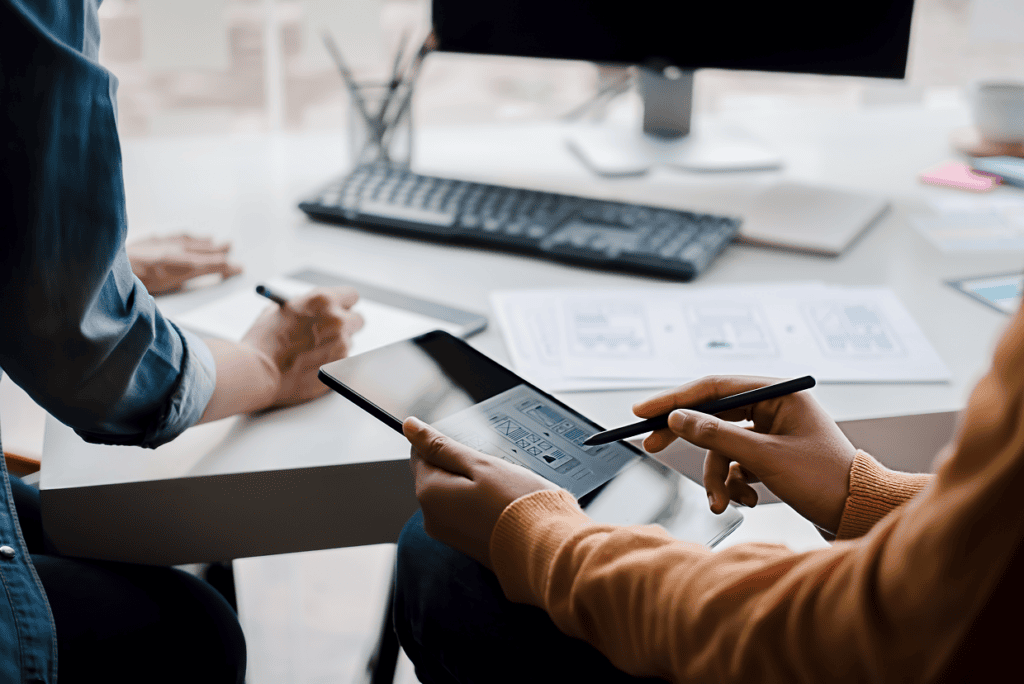 Hands holding a tablet and a pencil in front of a white desk with paper mockups, keyboard, and desktop monitor