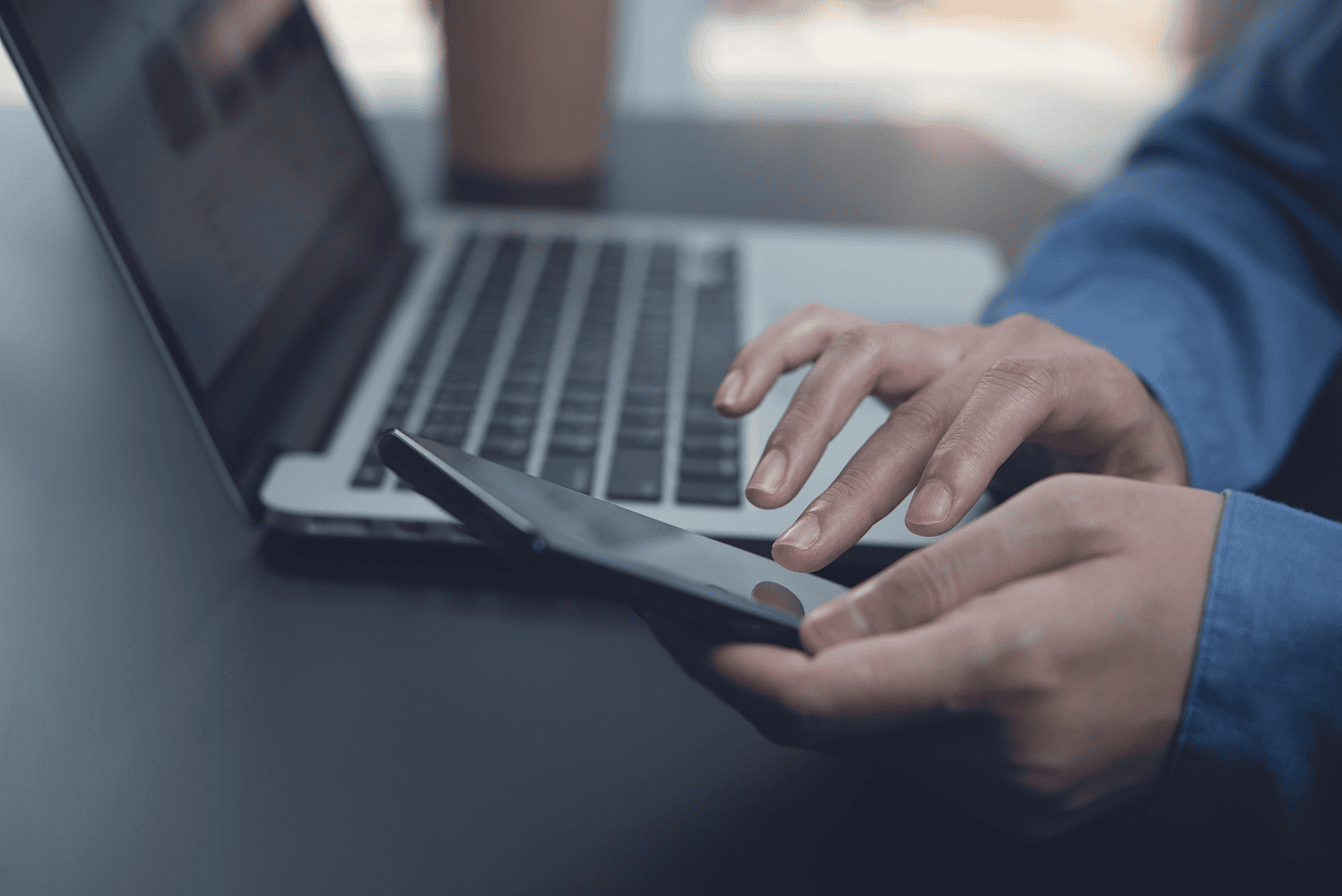 hand typing on a cell phone with a laptop on in the background
