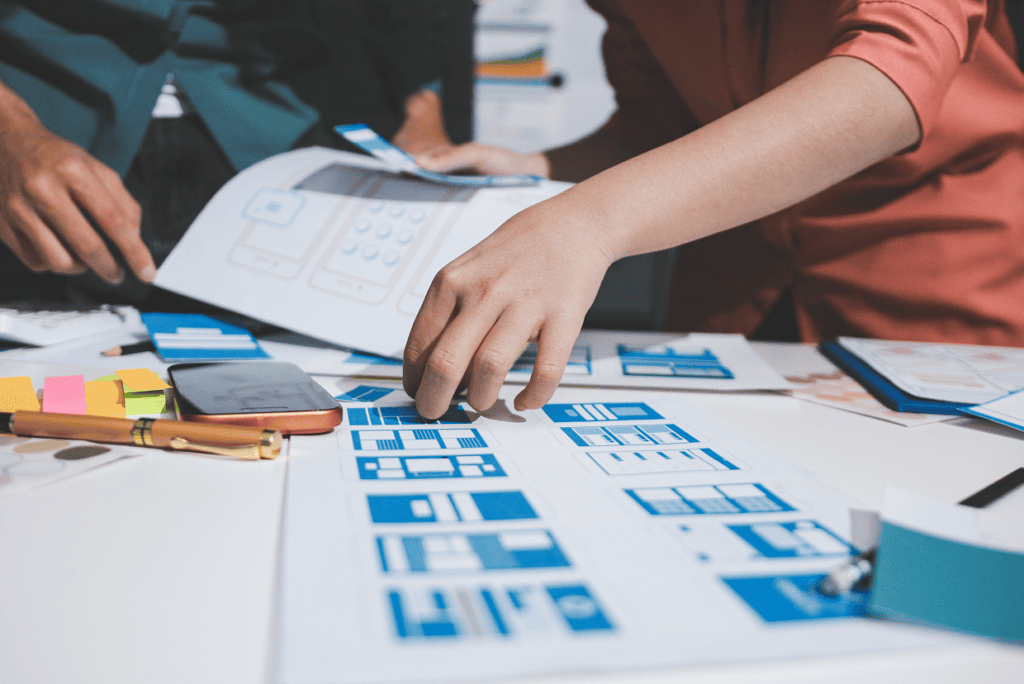 Desk with multiple blue paper mockups, post it notes, phone , pen, charts with 2 individuals
