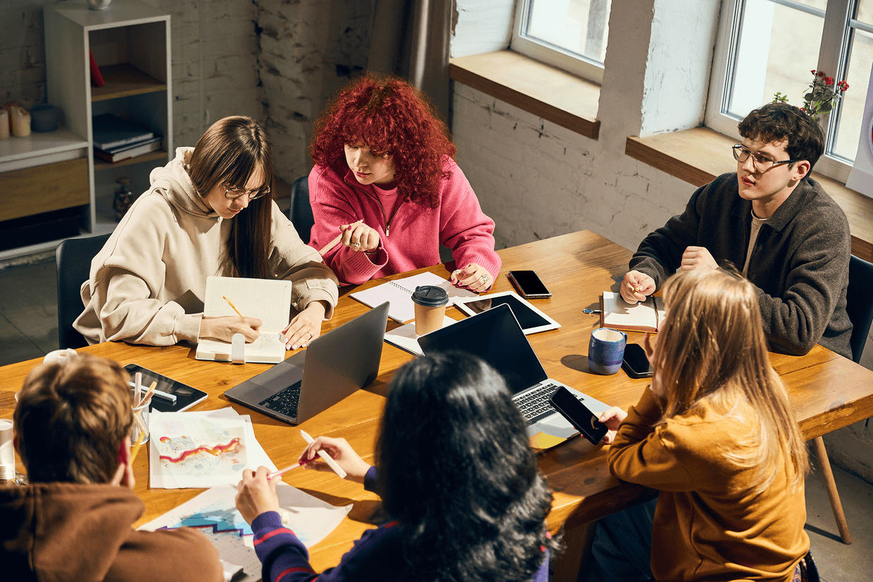 A group of young adults gathered around a table with coffee cups, laptop, tablets, charts and phone